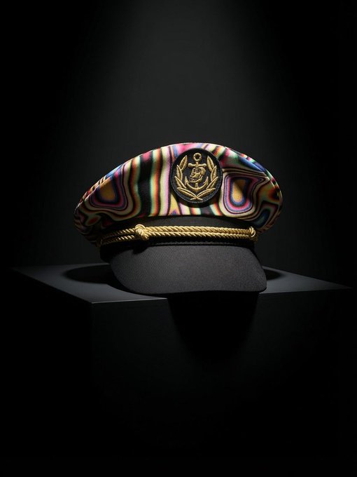 The image features a hat with a gold chain strap, which has a black band around its base. It appears to be a military-style hat or a captain's hat, as indicated by the gold chain strap. The hat is placed on a black stand against a dark background, creating a striking contrast that highlights the details of the hat and its design.