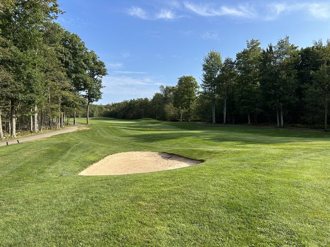 A golf course scene shows a sand bunker in the foreground with a green fairway extending into the distance, surrounded by trees under a blue sky.