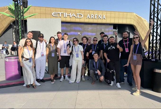 A group of people pose for a photo in front of the Etihad Arena, smiling and giving thumbs up, suggesting they attended an event or conference at the venue.