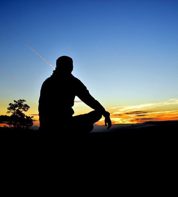 A silhouette of a person sitting cross-legged at sunset, with a tree visible on the left and a contrail in the sky.