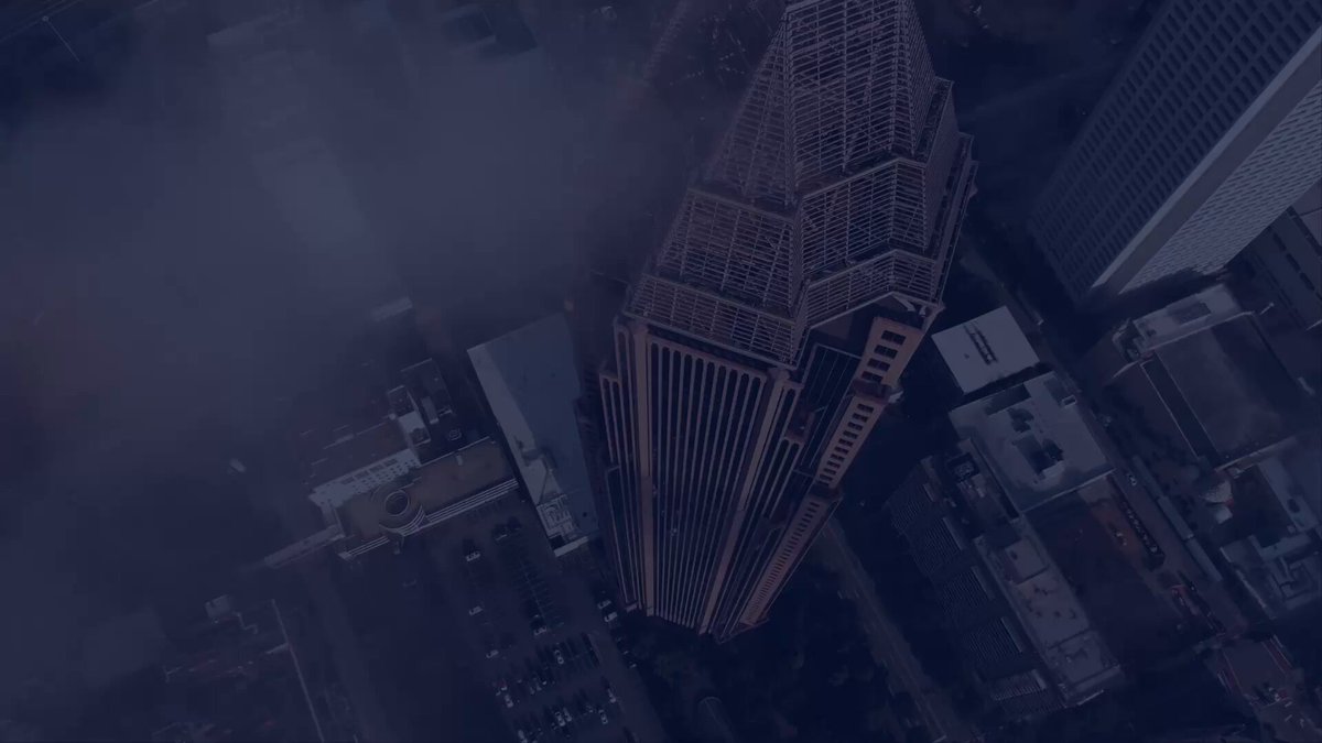 The image presents an aerial view of a city with tall buildings surrounding a dense urban landscape. One of these towering structures appears to be a skyscraper, possibly located near the top right corner of the frame. The buildings are densely packed together, indicating a dense urban environment. Below this prominent building, there is another smaller structure that seems out of focus. A large body of water can also be seen in the middle left portion of the image, adding to the overall scenery and complexity of the city's landscape.