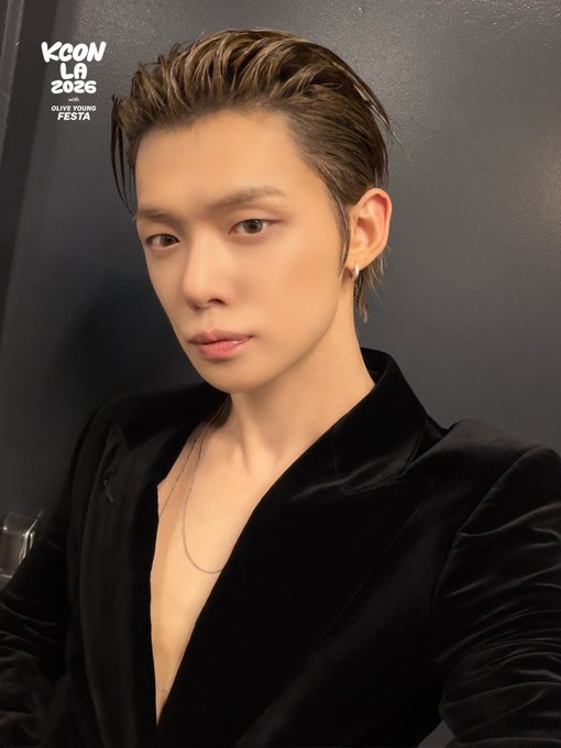 A selfie of Yeonjun from TXT taken backstage at KCON LA 2026, featuring the event logo "KCON LA 2026 Olive Young Festa" in the top left corner. He is wearing a black velvet deep-V blazer and posing with a confident expression against a dark background.