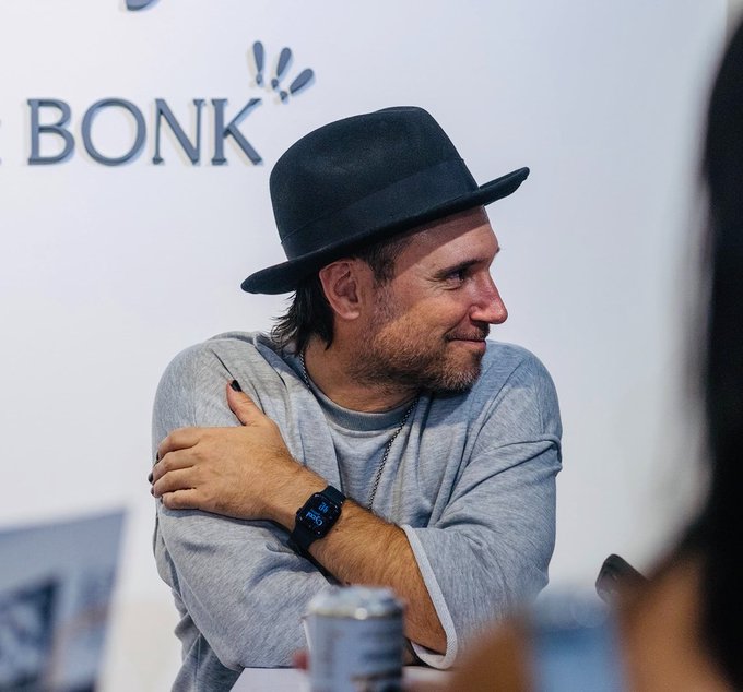 A man wearing a black fedora and gray long-sleeve shirt smiles while seated at what appears to be a panel or interview, with the "BONK" logo visible on the backdrop behind him, suggesting his connection to the Bonk Inu project mentioned in the post. A smartwatch is visible on his wrist as he crosses his arms in a relaxed pose, capturing a candid moment of reflection as he transitions from his role with Bonk to leading Cyclo Gallery.