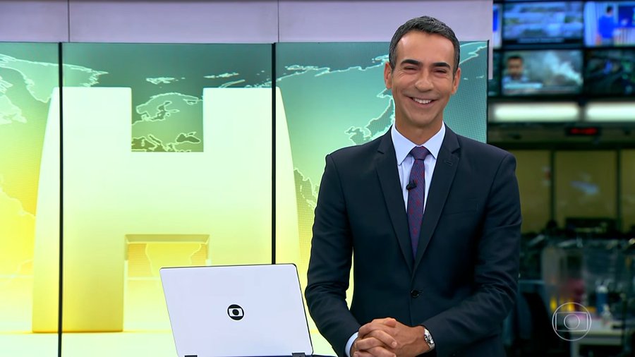 A male news anchor in a dark suit and patterned tie smiles while standing behind a laptop in a broadcast studio, with a large green "H" graphic and world map displayed on screens behind him. The accompanying post, referencing cycles of success and unseen failure, contrasts with the anchor's bright, polished on-air demeanor, suggesting that despite his confident smile, setbacks occur out of view of the audience.