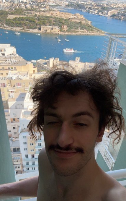 A shirtless man with curly hair winks at the camera from a high-rise balcony overlooking a Mediterranean harbor, likely in Malta, with boats dotting the blue waters and historic buildings lining the coast — a relaxed "GM" selfie asking for 500 comments.