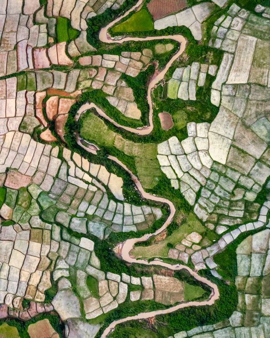 An aerial view shows a winding river snaking through a patchwork of green agricultural fields and rice paddies.