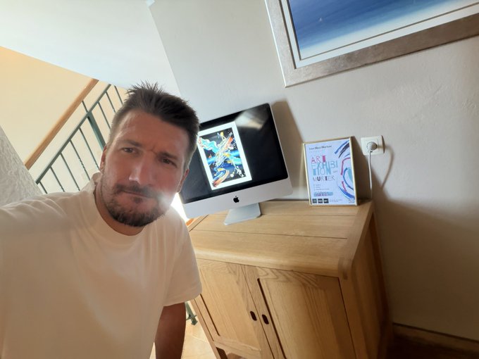 A man with a beard takes a selfie in a room with an iMac on a wooden cabinet, with a book or device displaying 'KUBERA' visible on the desk.