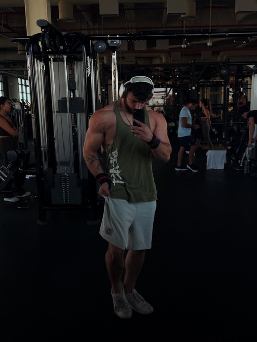 A muscular man in an olive green tank top and white shorts takes a mirror selfie in a gym, wearing headphones and looking down at his phone, with cable machines and other gym-goers visible in the dimly lit background. The casual "GM" (good morning) post pairs with his early gym session vibe, capturing a quiet moment of dedication before the day's workout.