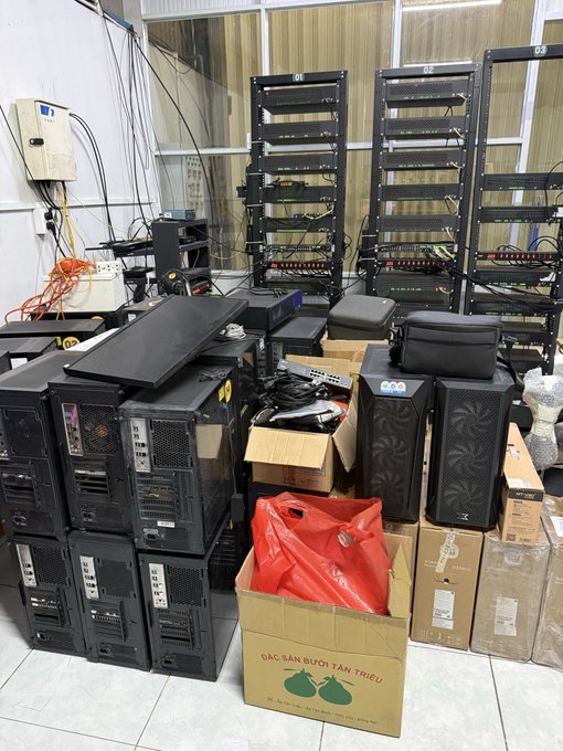 A cluttered storage room or shop contains stacked black computer towers, network racks, various boxes, and a cardboard box labeled 'CÁC SẢN PHẨM TỪ TÂN TRIỀU' with green citrus illustrations.