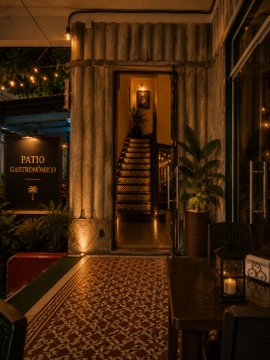 The image shows a restaurant entrance with a tiled floor and columns, which appears to be an old building. The entrance has a sign that says "PATIO GASTRONOMOCCO", indicating that this location is a restaurant or bar. There are two chairs visible in the scene - one near the entrance of the establishment and another further back inside the restaurant. A potted plant can also be seen, adding some greenery to the space. The image also contains text about Bitcoin meetups taking place at this location, suggesting that it is a venue for such events or gatherings related to cryptocurrency.