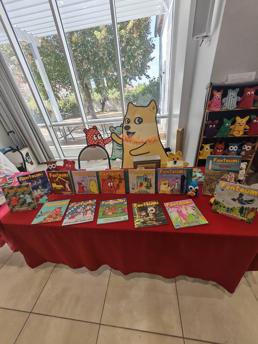 A display table covered with a red cloth showcases numerous 'Fant Tsum!' books and a cardboard Shiba Inu figure, alongside plush toys and a cat stand.