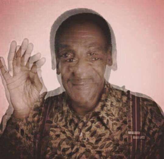 An older man in a patterned shirt smiles and raises his right hand in an ‘OK’ gesture against a plain wall.