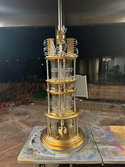 The image features a large gold metal tower with multiple levels of glass windows, giving it an elegant appearance. The tower is situated outdoors, possibly under a light source or at night, adding to its majestic presence. In front of the tower, there's a table and chairs, suggesting that this might be a gathering place for people to enjoy the view or engage in conversation.