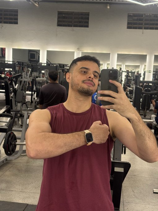 A man taking a mirror selfie in a gym, wearing a maroon sleeveless shirt, flexing his bicep while holding a smartphone, with gym equipment visible in the background.