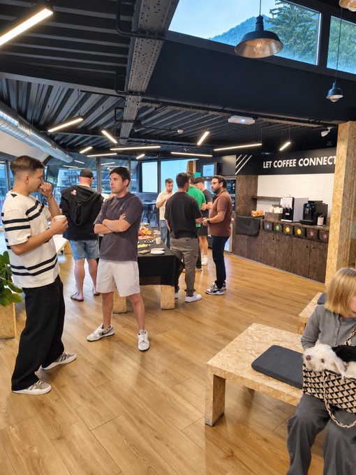 An indoor scene of a modern coffee shop or networking space where several people are standing and talking, with a sign reading 'LET COFFEE CONNECT' on the wall.