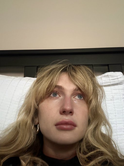 A young woman with wavy blonde hair lies on a bed with a melancholic, teary-eyed expression, humorously capturing the disappointment of being the only one whose "bags" aren't performing well in a trading context. Her sad gaze upward perfectly embodies the frustration of seeing others succeed while her own investments lag behind.