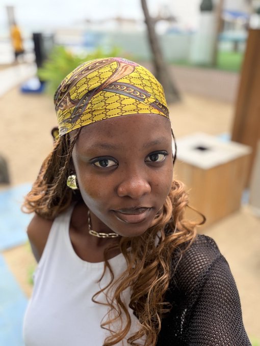 A close-up portrait shows a woman wearing a yellow and brown patterned headwrap and a white top, with a beach scene visible in the background.