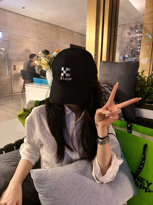 A young woman wearing a white shirt and a black 'X Layer' cap makes a peace sign while sitting on a gray sofa with pillows and a green shopping bag nearby.