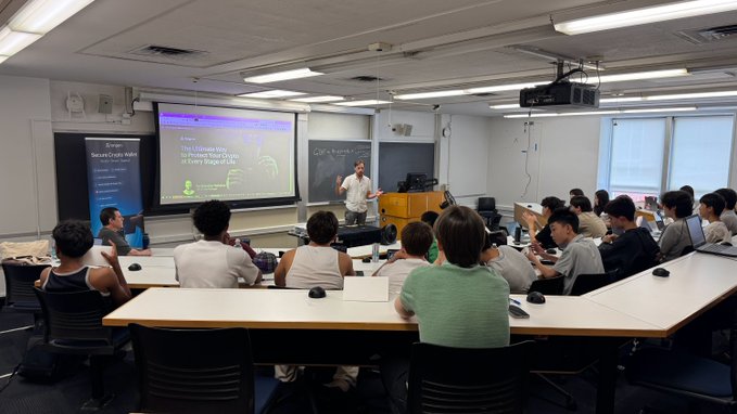 A presenter from Tangem stands at the front of a Columbia University classroom, engaging with high school students attending the Pre-College Program's "Blockchain, Cryptocurrencies, AI, and Beyond" course. A projection screen behind him displays the Tangem logo alongside the message "The Lifetime Way to Protect Your Crypto in Every Shape or Size," reflecting the brand's involvement in educating future builders about crypto security.