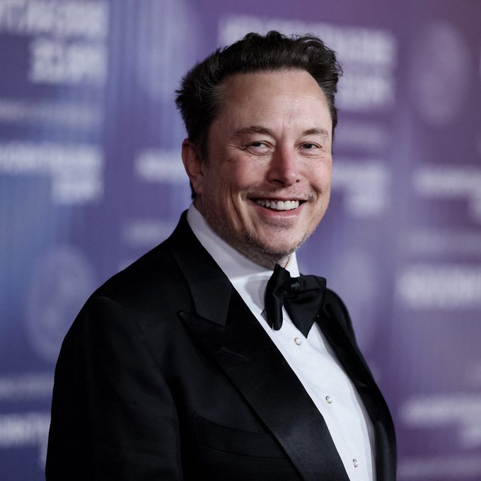 Elon Musk smiles while wearing a black tuxedo with a bow tie, photographed against a purple backdrop with blurred text.