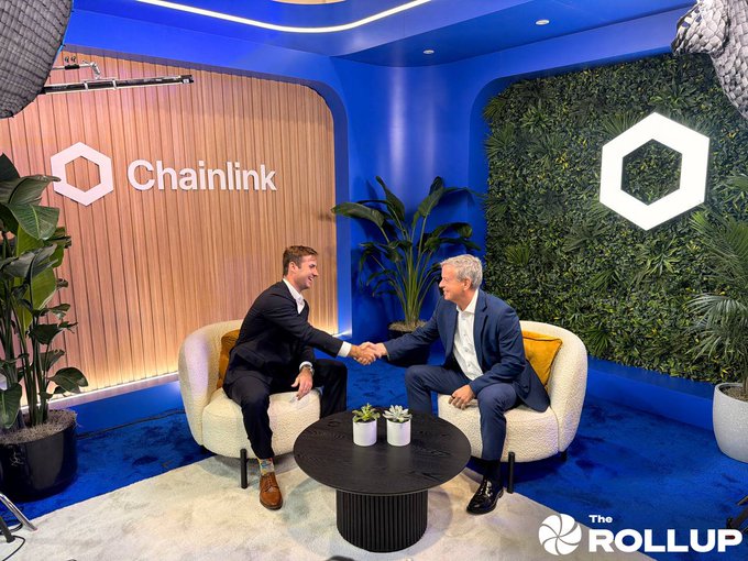 Two men in business suits shake hands while seated in a modern studio setting with a blue ceiling and a 'Chainlink' logo on a wooden wall, branded with 'The Rollup' logo.