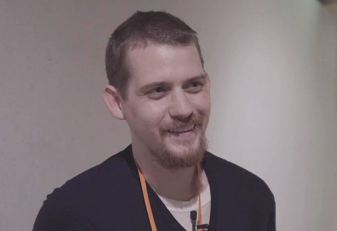 A man with short brown hair, a goatee, and a friendly smile appears to be speaking at an event, wearing a dark sweater and an orange lanyard with a microphone clipped nearby, likely depicting Luke Dashjr as referenced in the post about his claim that exchanges sell "fake Bitcoin."