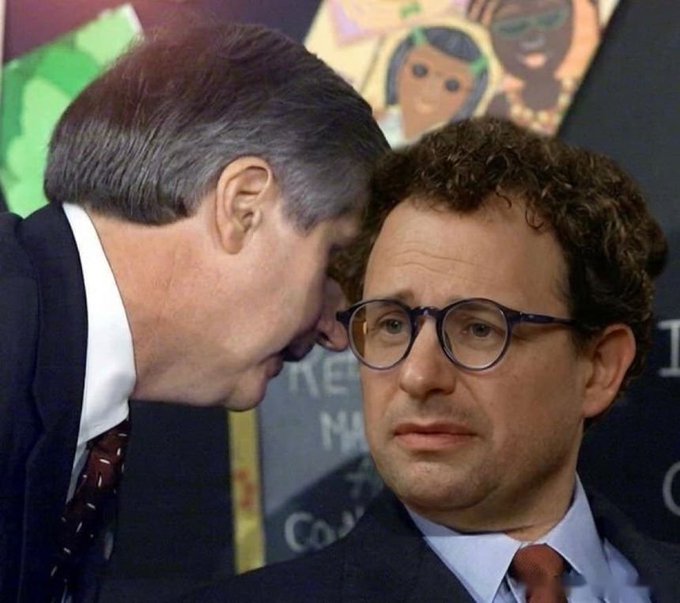 Two men in suits are shown in a close-up, with one leaning in to whisper into the ear of a wide-eyed, bespectacled man who looks uncomfortable.