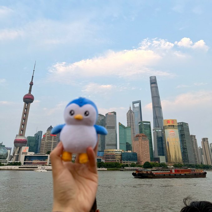 A hand holding up a small blue and white penguin plush toy in front of the Shanghai skyline, including the Oriental Pearl Tower and other Pudong skyscrapers along the Huangpu River.