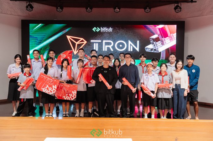 A group of students and adults posing on a stage with a 'TRON' logo on the digital backdrop, alongside 'bitkub blockchain' branding, holding red Bitkub promotional items.
