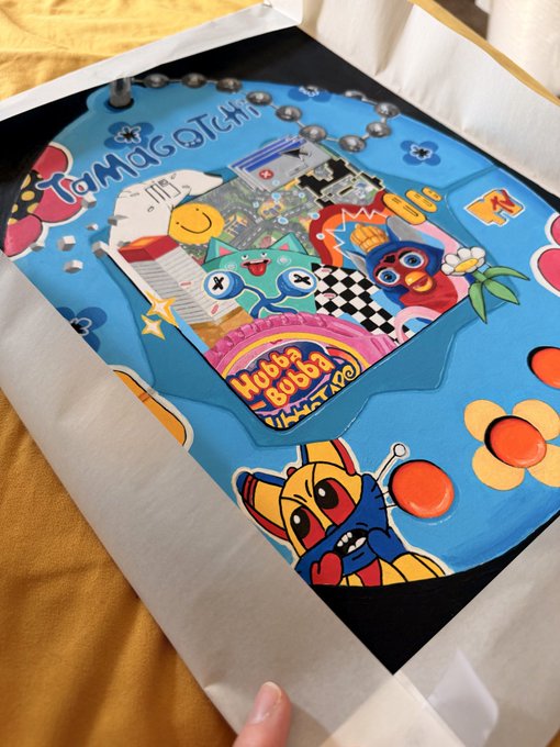 A Tamagotchi-themed artwork or product features various colorful characters, a 'Hubba Bubba' logo, flowers, and a smiley face, partially wrapped in white paper on a yellow surface.