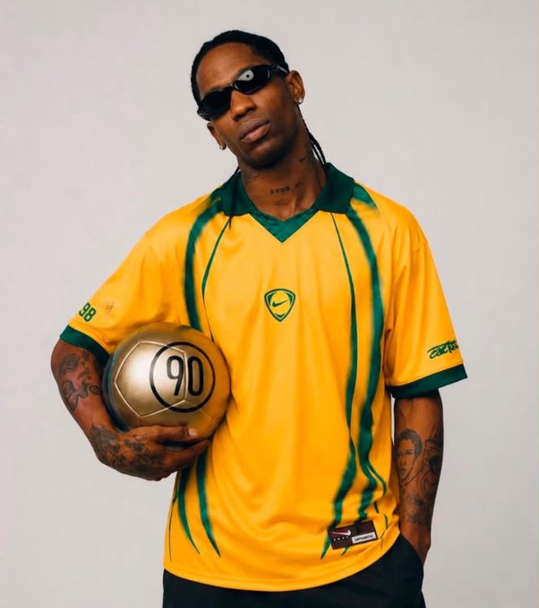A photo of Travis Scott wearing a yellow and green Brazil-style soccer jersey with the number '90' on a gold ball he's holding, posing against a plain white background.