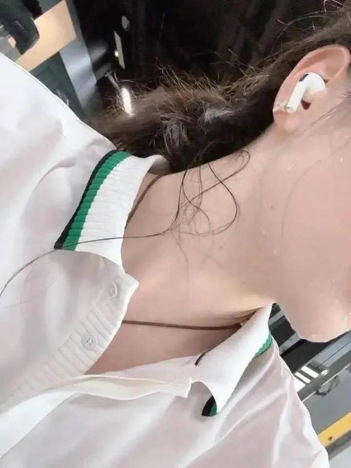 The image features a person wearing a white shirt with green trim, who has an earbud in their ear. The earbud appears to be connected to a device or gadget that is not visible in the photo. The background of the image shows a chair and a desk, suggesting that this might be taking place in a workspace or office setting.