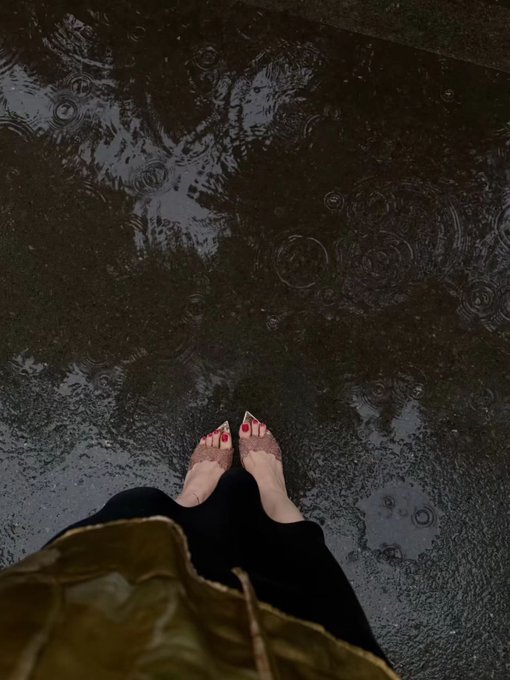 Overhead photograph showing a person's feet in pink studded sandals standing in a puddle on wet pavement, with rain ripples visible on the water's surface.