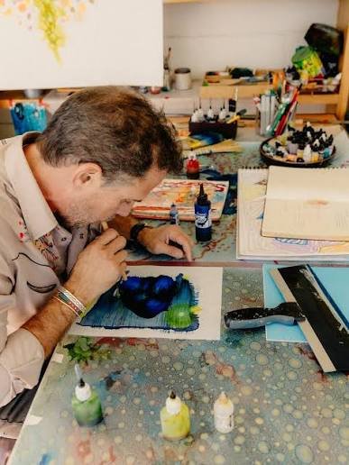A man with short gray hair and a beard works on a colorful watercolor painting in his art studio, surrounded by ink bottles, brushes, and artwork.