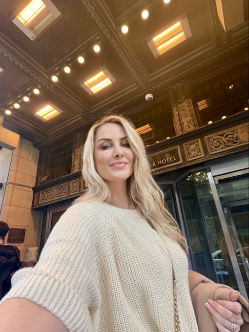 A low-angle selfie of a blonde woman wearing a cream knit sweater, standing outside an ornate building with a sign partially reading 'ER HOTEL'.