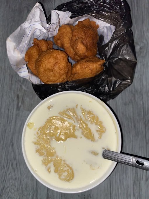 A bowl of creamy soup or porridge next to a basket of fried chicken pieces in a plastic bag, with a metal spoon resting in the bowl.