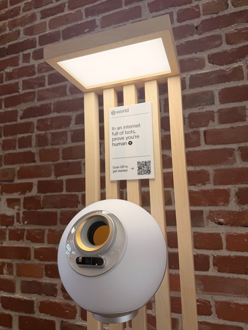 A sphere-shaped biometric "World" ID verification device sits mounted beneath a light fixture against a brick wall, accompanied by a sign reading "In an internet full of bots, prove you're human" with a QR code to scan for getting started—directly illustrating the post's message about keeping the internet for humans.