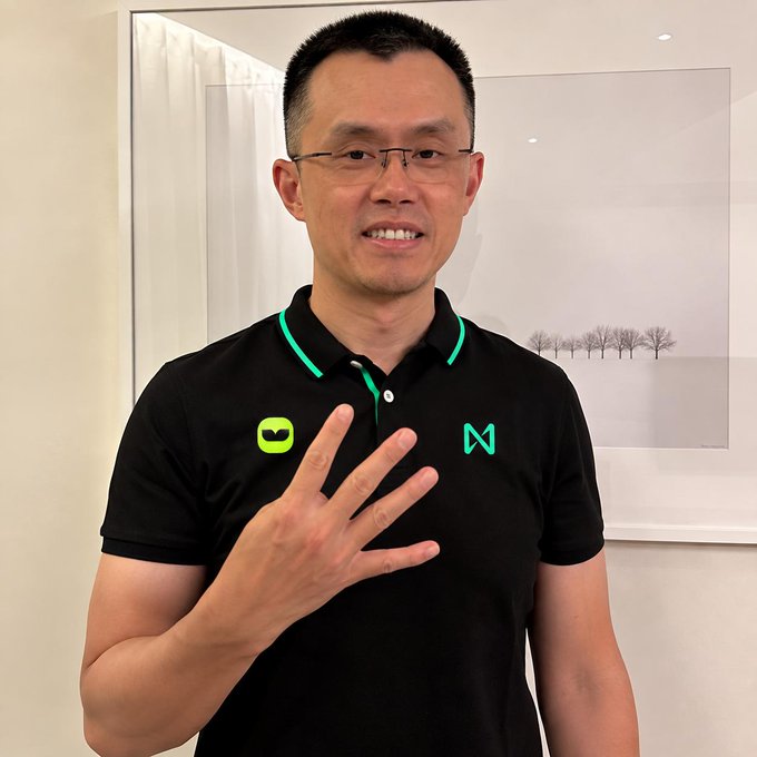 A man with glasses holding up four fingers, wearing a black polo shirt with green logos including an 'M' and a superhero mask icon.