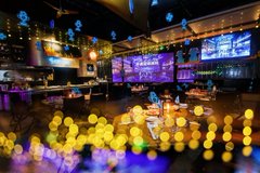 A dimly lit bar or restaurant interior with colorful neon lighting, candles glowing on tables, and a string of hanging lights creating a moody ambiance.