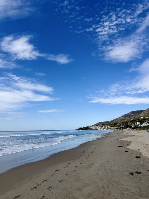 A serene coastal scene showing a wide sandy beach with gentle waves under a bright blue sky filled with scattered white clouds, with mountains and a small town visible along the shoreline in the distance.