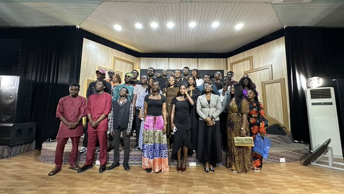 A group of around twenty people pose together for a photo on a carpeted floor in front of a stage with black curtains and an air conditioning unit.
