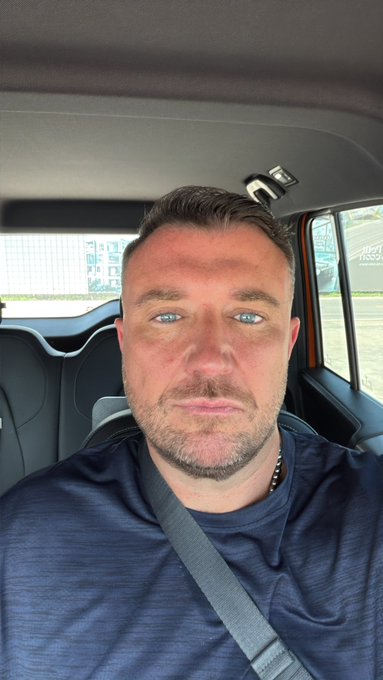 A selfie of a man with short hair and blue eyes, wearing a dark blue shirt and seatbelt, sitting inside a car.
