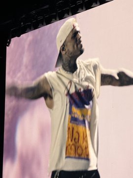 A performer, appearing to be Chris Brown, on stage wearing a white tank top with colorful graphics and a white cap, arms outstretched in front of a large screen.