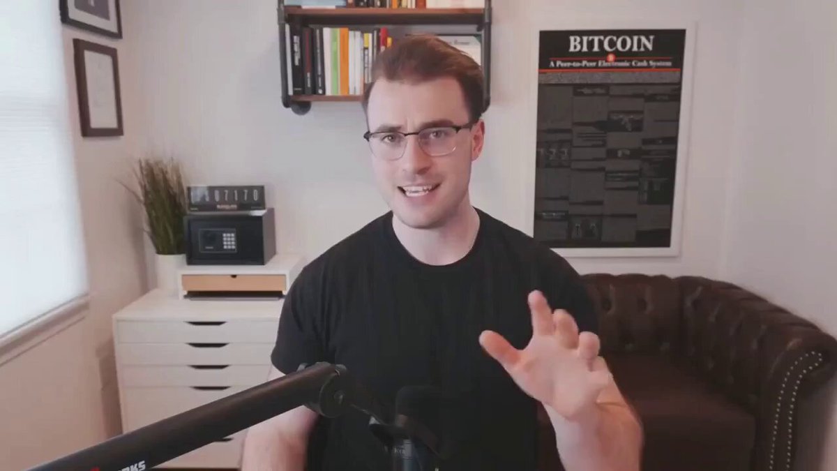 The image shows a man wearing glasses sitting behind a microphone, with a couch visible in the background. The man appears to be giving a speech or presentation, possibly related to the sale of Bitcoin. He is holding up his hand as if he's about to make a gesture or point at something. The presence of books and a potted plant in the room suggests that it might be an office or workspace setting where strategy discussions are held.