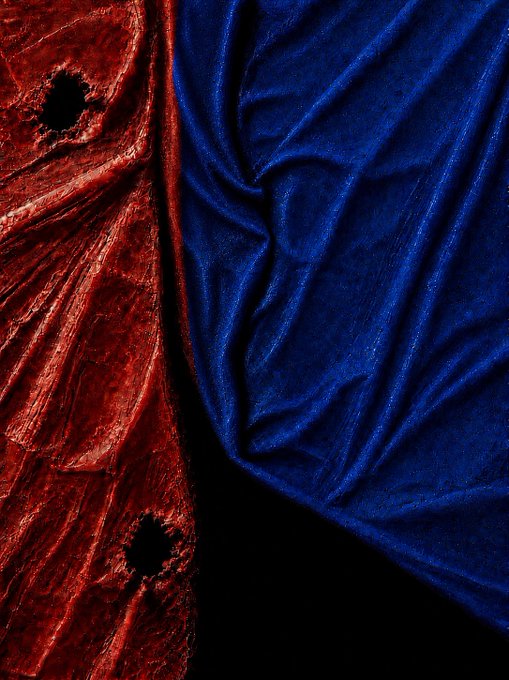 An artistic close-up shows crumpled red velvet fabric with two small holes on the left and draped, lustrous blue velvet fabric on the right, set against a black background.