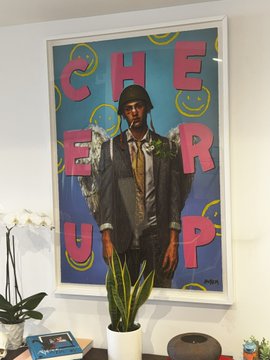 A framed pop art poster features a man in a suit and hat holding flowers, with large pink text spelling 'CHEER UP' and yellow smiley face patterns on a blue background.