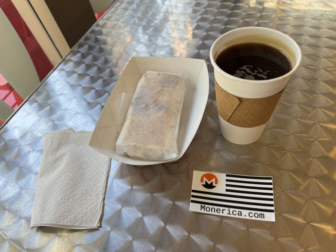 A coffee cup with a brown sleeve sits on a metal table next to a paper tray holding what appears to be a wrapped pastry, with a business card displaying a Monero logo and "Monerica.com" placed in the foreground.