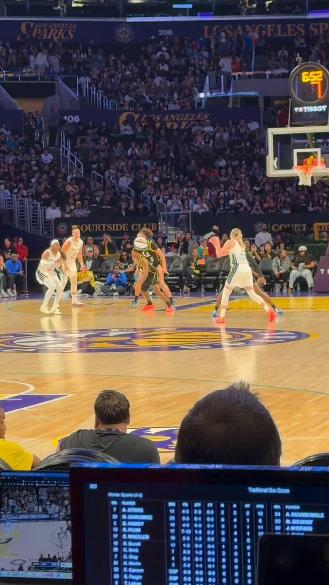 The image shows a basketball game taking place, with players actively engaged in the match. A man can be seen sitting down while watching the game from his laptop screen. The laptop screen displays information about the game, including the score of the game being played at the moment and the names of the teams playing against each other. This suggests that the viewer is interested in following the progress of the game or learning more about it.