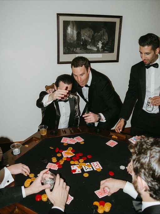 A group of men in tuxedos playing poker around an octagonal table covered in chips and cards, with a classical framed painting on the wall behind them.