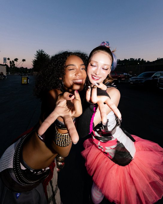 Two smiling women pose playfully for the camera, with the woman on the right wearing a colorful outfit featuring a pink tulle skirt and a floral headpiece, set against a dusky outdoor backdrop with parked cars. The candid, joyful pose captures the fun, carefree atmosphere likely associated with the "Head in the Clouds" event mentioned in the caption.